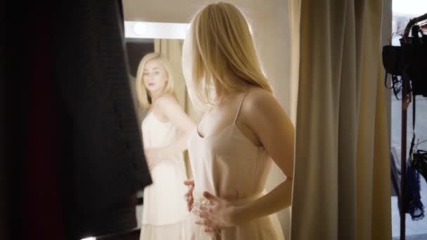 Underbar blond vit kvinna i beige klänning tittar i stor spegel i mode butik. Vacker ung shoppare provar kläder i omklädningsrummet. Skönhet, mode, shopping, stil. Långsam. — Stockvideo