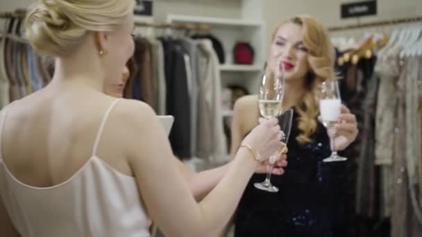 Tre positiva vita kvinnor klinkar glas med champagne i modebutiken. Glada rika kvinnliga vänner firar inköp i köpcentret. Rikedom, livsstil, konsumism. Långsam. — Stockvideo