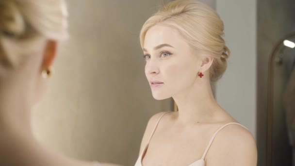 Close-up reflection of beautiful blond Caucasian woman with grey eyes looking at mirror and smiling. Attractive young girl posing indoors. Backstage, fashion, elegance. — Stock Video