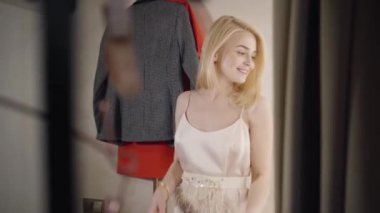 Soyunma odasında gülümseyen ve dönen mutlu beyaz kadın portresi. Moda mağazasında kıyafet seçen neşeli sarışın kadın. Yaşam tarzı, neşe, alışveriş.