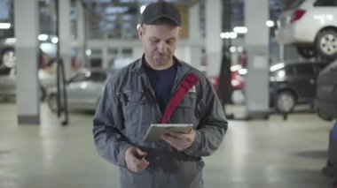 Otomobil tamircisine yaklaşan kamera tableti tutuyor ve arabanın anahtarlarını kameraya gösteriyor. Tebessüm eden beyaz bir adam atölyede poz veriyor. Otomotiv endüstrisi, otomobil bakımı, hizmet.