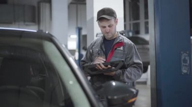 Ciddi bir beyaz bakım mühendisinin portresi dijital ekipmanlarla otomobili kontrol ediyor. İş kıyafetlerindeki yetişkin adam araba hasarını doğrulamak için bilgisayar kullanıyor. Tamirhane, benzin istasyonu..