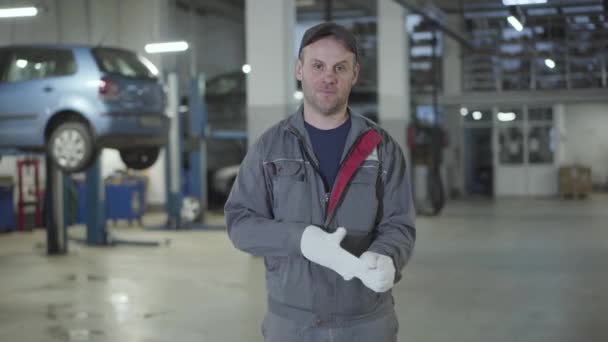 Portrait of young confident Caucasian man posing in auto repair shop. Professional male auto mechanic taking off working gloves and smiling at camera. Lifestyle, occupation, job. — Stock Video