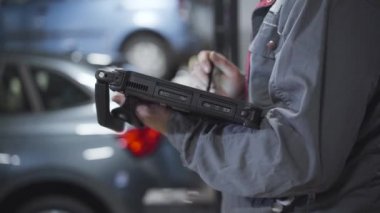 Tamir atölyesinde ekipmanla çalışan tanınmayan erkek oto tamircisinin yan görüntüsü. Benzin istasyonunda otomobilin arka planında bilgisayar tutan beyaz bir adam. İş, yaşam tarzı, endüstri.