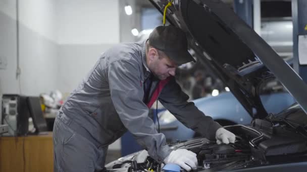 Ingeniero de mantenimiento caucásico profesional de pie en el capó abierto del coche y estimar los daños. Hombre serio en ropa de trabajo y guantes de protección fijación de rotura en taller de reparación de automóviles . — Vídeos de Stock