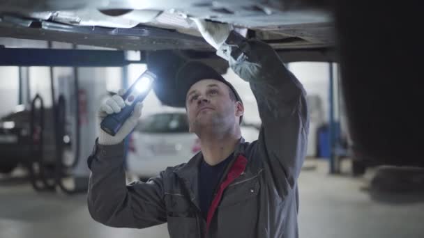Porträtt av vuxen vit underhållsingenjör belysning på bilens botten med ficklampa och kontrollera botten manuellt. professionell vuxen man auto mekaniker undersöka brott. — Stockvideo