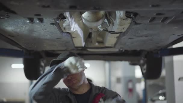 Hombre caucásico adulto limpiando la frente con la mano como tornillos de apriete en la parte inferior del coche. Retrato de mecánico de automóviles masculino profesional que trabaja en taller de reparación de automóviles. Estilo de vida, trabajo, industria automotriz . — Vídeo de stock