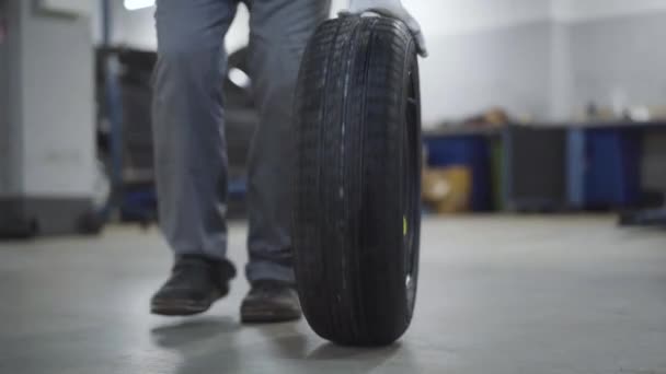 Unrecognizable worker rolling car wheel in repair shop. Man in workwear and protective gloves walking to camera. Automobile industry, tuning, insurance service station. — Stock Video