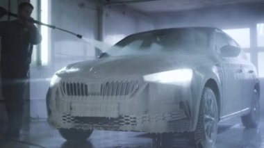 Araba yıkama servisinde farları yanan araba. İşçi, araç yüzeyinden köpüğü yüksek basınçlı spreyle temizliyor. Bakım, endüstri, iş.