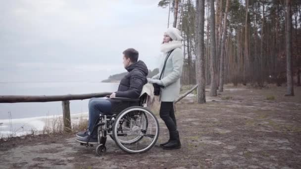 Mladá běloška drží vozík s postiženým chlapcem a mluví. Kamera pohybující se zleva doprava kolem handicapovaného muže a neplatná něha stojící v lese na břehu řeky. — Stock video