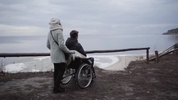 Back side view of woman standing with wheelchair with disabled boy in it. Caucasian crippled man and mother or invalid tender spending autumn day on river bank outdoors. Support, care, disability. — Stock Video