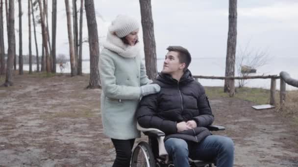 Portrait of smiling boy in wheelchair and adult woman talking and holding hands. Caucasian handicapped man enjoying time outdoors with mother or invalid tender. Lifestyle, happiness, disability. — Stock Video