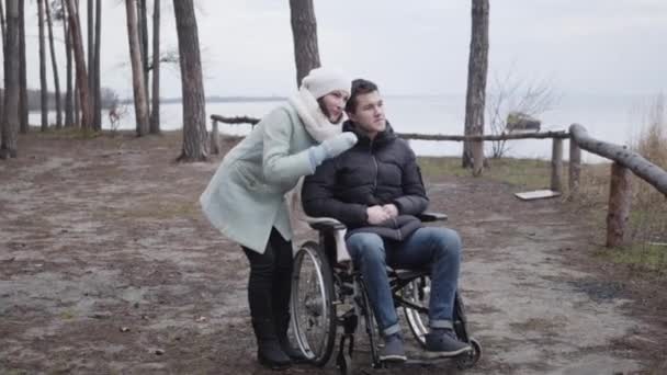 Brede foto van een glimlachende blanke vrouw die wegwijst en praat met een gehandicapte jongen in een rolstoel. Positieve invalide die de herfstdag doorbrengt met invalide persoon buiten in het bos op de oever van de rivier. — Stockvideo