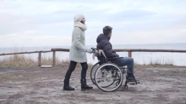 Nehir kıyısında engelli bir çocukla tekerlekli sandalyede oturan yetişkin beyaz kadın. Gülümseyen engelli bir adam ve güçlü bir kadın güz gününün tadını çıkarıyor..