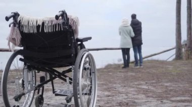 Arka planda iki bulanık insanın konuştuğu tekerlekli sandalyeye yakın çekim. Beyaz erkek ve kadın, sonbahar gününün tadını çıkarıyorlar. Engelli, felçli, sağlık hizmetleri.