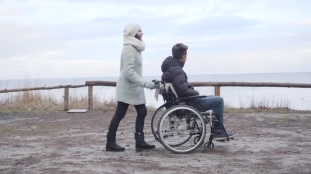 Vista laterale della donna caucasica adulta che rotola sedia a rotelle con ragazzo handicappato lungo la riva del fiume. Fotocamera seguente sorridente giovane uomo disabile e donna forte godendo di giornata autunnale all'aperto . — Video Stock