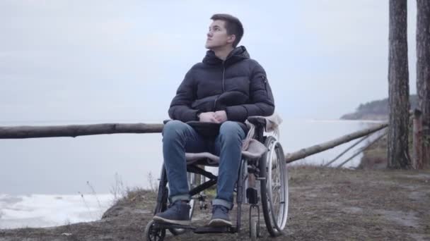 Brede opname van een bedachtzame blanke gehandicapte jongen in een rolstoel die buiten rondkijkt. De jonge brunette brengt de herfstdag alleen door op de oever van de rivier. Eenzaamheid, levensstijl, handicap. — Stockvideo