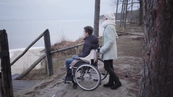 Zijaanzicht van vrolijke blanke gehandicapte jongen die praat met invalide teder en lachend. Portret van een kreupele jongeman in een rolstoel die geniet van vrije tijd buiten. Lifestyle, verzorging, hulp. — Stockvideo