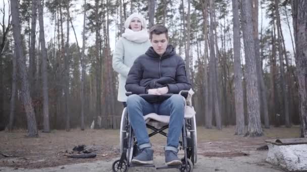 Volwassen blanke vrouw met een schouder van zielige gehandicapte jongen in een rolstoel en pratend. Moeder of invalide die de herfstdag doorbrengt met patiënt of zoon buiten in het bos. Lifestyle, vrije tijd, zorg. — Stockvideo