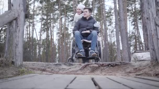 Portret van een glimlachende blanke kreupele man in een rolstoel die praat met een vrouw buiten in het bos. Jonge gehandicapte man die tijd doorbrengt met een invalide of moeder. Gehandicapten, kracht, levensstijl. — Stockvideo