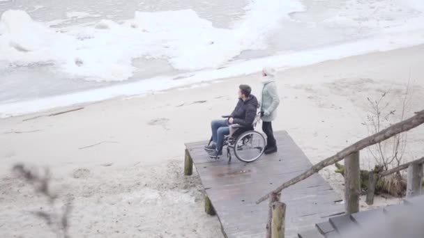 Blanke gehandicapte jongen in rolstoel en invalide tender die op de pier staat en kijkt naar bevroren rivier. Jonge gehandicapte man geniet van de herfstdag met moeder buiten. Hulp, verzorging, levensstijl. — Stockvideo