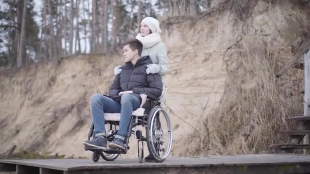 Overstuur blanke jongen zit in een rolstoel met moeder of invalide teder. Verdrietige kreupele man die de herfstdag buiten door brengt in het bos. Concept van sociale kwesties, medische problemen, levensstijl, handicap. — Stockvideo