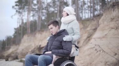 Tekerlekli sandalyedeki yakışıklı, engelli çocuk dışarı ve aşağıya bakıyor. Ciddi bir beyaz kadın omuzlarını tutuyor ve düşünüyor. Felç, sakatlık, yaşam tarzı, rehabilitasyon.