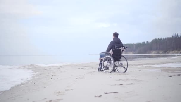 Weitschussbild eines jungen behinderten Kaukasiers im Rollstuhl am Ufer des Herbstflusses. Brünetter behinderter Junge, der sich im Freien ausruht. Einsamkeit, Behinderung, Lähmung, Lebensstil.