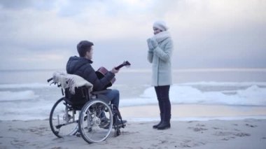 Gülümseyen genç anne el çırpıyor ve engelli oğlunu tekerlekli sandalyede kucaklıyor. Kumral adam deniz kıyısında ana baba için gitar çalıyor. Destek, rehabilitasyon, bakım, yaşam tarzı, hobi.