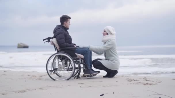 Tekerlekli sandalyedeki bir çocuğun önünde oturan ve konuşan genç bir kadın. Deniz kıyısında ya da nehir kıyısında engelli bir genci destekleyen anne ya da yatalak ihale. Neşe, yaşam tarzı, yardım. — Stok video