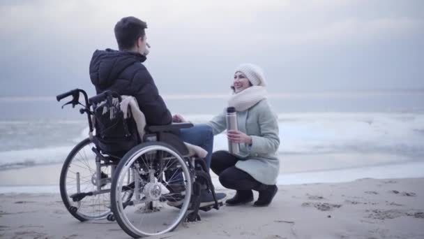 Νεαρό ανάπηρο λευκό αγόρι σε αναπηρική καρέκλα πίνοντας τσάι στη θάλασσα και μιλώντας με όμορφη γυναίκα κάθεται σε hunkers. Νεαρός με ειδικές ανάγκες περνάει την ημέρα του φθινοπώρου με θετική προσφορά. — Αρχείο Βίντεο