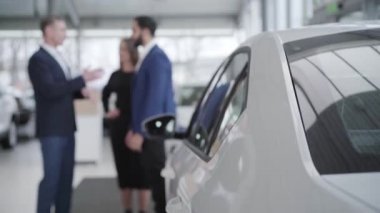 Araba galerisinde konuşan bulanık insanlar, ön planda duran yeni beyaz otomobil. Orta Doğulu sakallı bir adam ve beyaz bir kadın galeride tüccarla alışverişi tartışıyor..