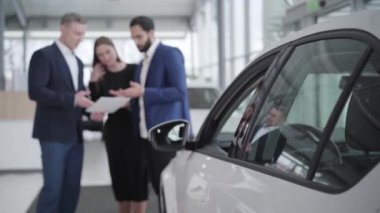 Bulanık araba satıcısı sergi salonundaki çok ırklı çifte kitapçığı gösteriyor. Orta Doğulu genç bir adam ve beyaz bir kadın galeride otomobil seçiyor. İş, başarı, yaşam tarzı, endüstri.