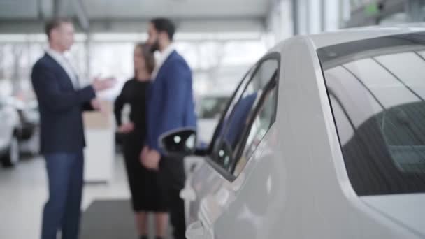 Pozitivně rozmazaní lidé mluví v prodejně aut, v popředí stojí nový bílý automobil. Vousatý muž ze Středního východu a běloška diskutují o nákupu s obchodníkem v showroomu. — Stock video