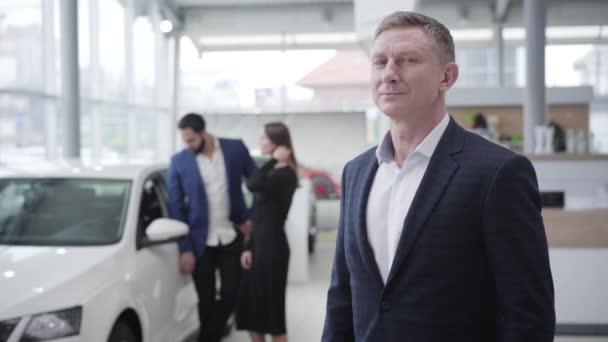 Confident Caucasian car dealer looking back at happy multiracial couple opening new white car and showing keys at camera. Smiling man posing in dealership. Showroom. business, lifestyle. Middle shot. — Stock Video