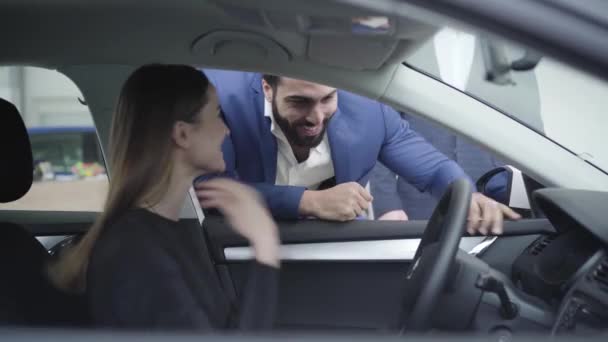 Joven hombre guapo de Oriente Medio hablando con una hermosa mujer caucásica sentada en el asiento del conductor. Exitoso hombre de negocios que compra automóviles para su novia o esposa en el concesionario. Industria automovilística, alegría . — Vídeos de Stock