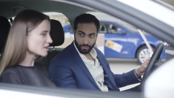 Retrato de belo homem do Oriente Médio sentado no carro com a esposa caucasiana como comerciante fechando portas. Empresário bem sucedido positivo escolher veículo com cônjuge na concessionária. Negócios, estilo de vida . — Vídeo de Stock