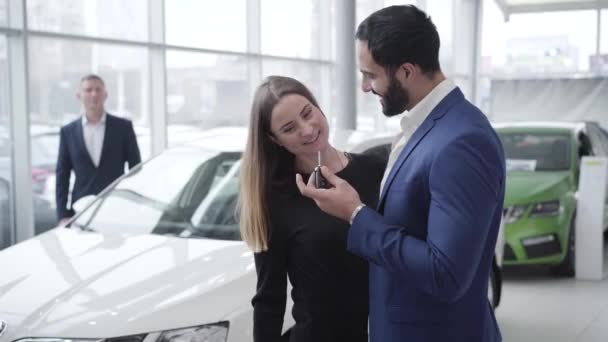 Gazdag, sikeres közel-keleti férfi ad kocsikulcsot a gyönyörű kaukázusi nőnek a bemutatóteremben. A boldog fiatal pár kamerába néz és mosolyog a kereskedésben. Luxus, életmód, vagyon. — Stock videók