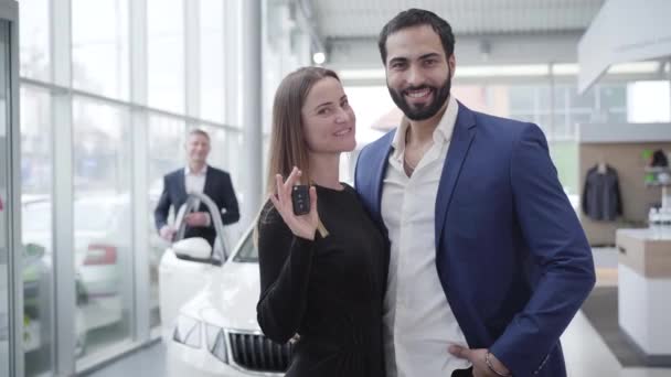 Junge wohlhabende multiethnische Familie posiert im Autohaus mit Autoschlüsseln. Hübsche kaukasische Ehefrau und hübscher Mann aus dem Nahen Osten, der in die Kamera schaut und lächelt. Lifestyle, Luxus. — Stockvideo