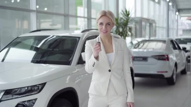 Joven rubia confiada mostrando las llaves del coche en la cámara y sonriendo. Mujer de negocios en traje blanco comprando vehículo en concesionario. Riqueza, lujo, estilo de vida, éxito . — Vídeos de Stock