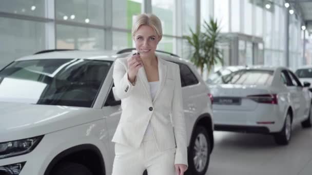Jonge zelfverzekerde dame draait naar de camera met autosleutels en glimlachen. Succesvolle blanke vrouw op zoek naar witte nieuwe auto staan in de showroom. Zakenvrouw koopt auto in dealerovereenkomst. — Stockvideo