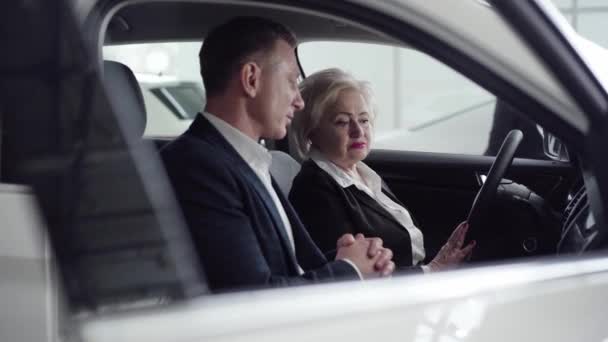 Egy gazdag fehér nő ül az autóban egy dílerrel. Professzionális kereskedő beszél gazdag ügyféllel a kereskedésben. Autóipar, üzlet, luxus, életmód. — Stock videók