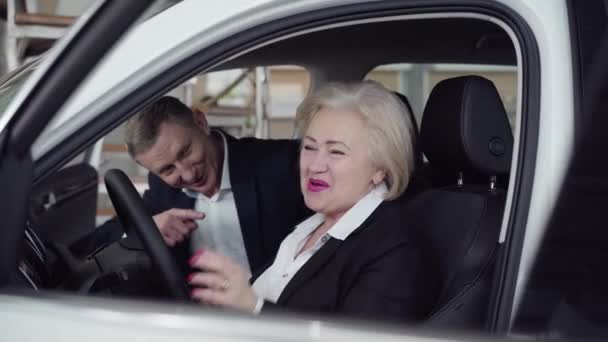Egy magabiztos fehér nő, aki beköti a biztonsági övet egy új autóban a bemutatóteremben. Pozitív gazdag ügyfél vizsgálja jármű kereskedő a kereskedésben. Életmód, üzlet, autóipar. — Stock videók