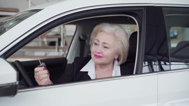 Portrét šťastné starší bělošky držící klíčky od auta, dívající se do kamery a usmívající se. Úspěšná blonďatá obchodnice kupující nové auto v prodejně. Luxus, obchod, životní styl, bohatství. — Stock video
