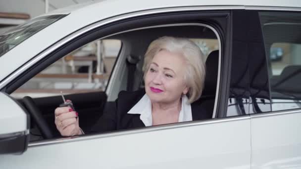 Retrato de una mujer caucásica mayor feliz sosteniendo las llaves del coche, mirando a la cámara y sonriendo. Exitosa empresaria rubia comprando un automóvil nuevo en el concesionario. Lujo, negocios, estilo de vida, riqueza . — Vídeo de stock