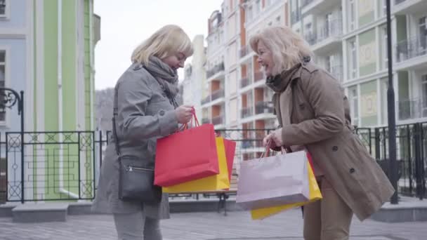 Veselé starší ženy se chlubí nákupy na City Street. Kavkazští důchodci středního věku si po nakupování venku povídají a smějí se. Štěstí, shopaholismus, životní styl, odchod do důchodu. — Stock video