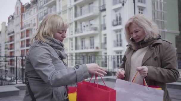 Pozitív középkorú kaukázusi nők portréja, akik a városi utcai vásárlásokról beszélgetnek. Két vidám aktív nyugdíjas bevásárlószatyorral, akik az őszi napot a szabadban töltik. Életmód, öröm, boldogság, gazdagság. — Stock videók