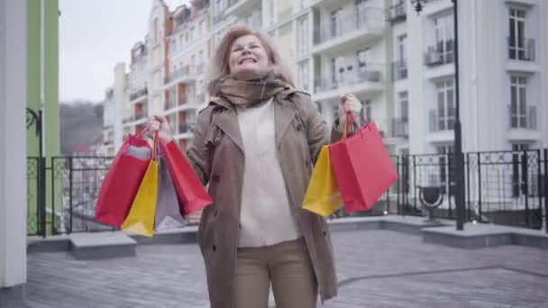 Porträt einer zufriedenen Seniorin, die mit Einkaufstüten auf der Straße dreht und tanzt. Ältere kaukasische Frau genießt den Herbstverkaufstag mit Einkäufen im Freien. Lebensstil, Freude, Wohlstand. — Stockvideo