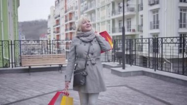 Pozitif kıdemli beyaz kadın alışveriş torbalarıyla şehir caddesinde duruyor ve gülümsüyor. Satın almaktan memnun, mutlu, sarışın bir kadının portresi. Alışveriş, yaşam tarzı, keyif..