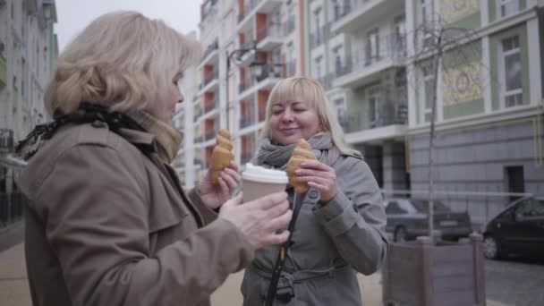 Старшие белые женщины пьют кофе и едят круассаны на городской улице. Счастливые активные пенсионеры отдыхают на свежем воздухе. Стиль жизни, досуг, пенсия, счастье . — стоковое видео